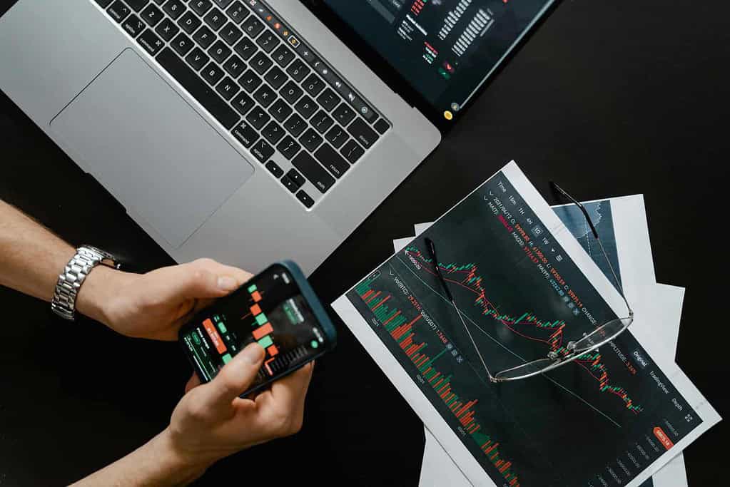 Investor reviewing stock charts and financial data on a mobile app while cross-checking with a laptop.